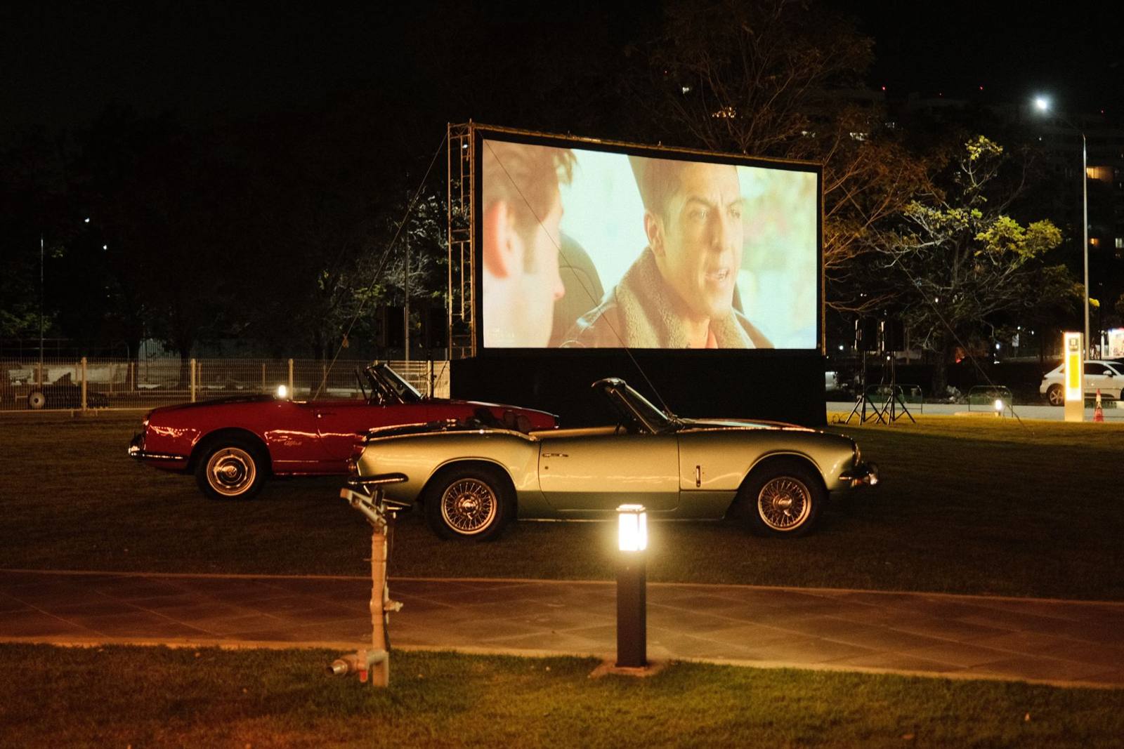 Two roadsters parked in front of a drive-in movie screen at night